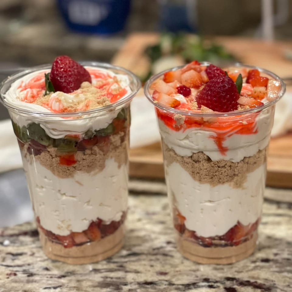 Two layered desserts in clear cups with strawberries, whipped cream, and cookie crumbles.