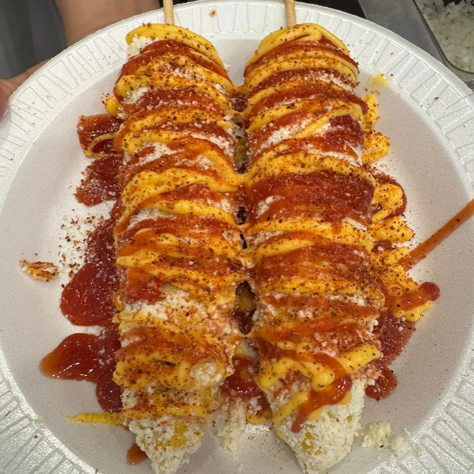 Two elotes, Mexican street corn, on a white plate, topped with cheese, sauce, and chili powder.